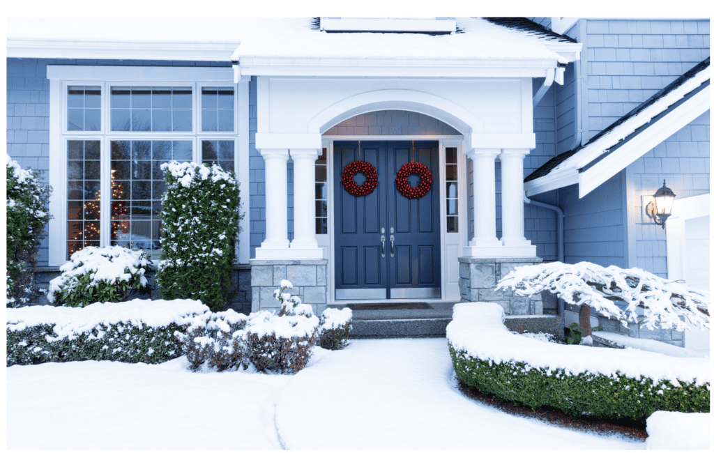 Snowy blue house with a double door entrance decorated with wreaths, and snow-covered bushes.