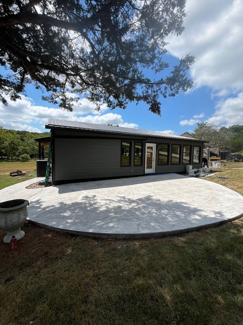 Small modern building with dark siding and large windows beside a round concrete patio under a tree-filled sky