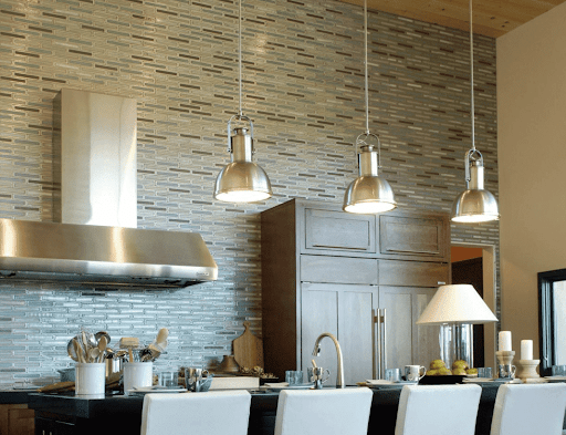 Modern kitchen with stainless steel appliances and pendant lights. Gray and beige tiled backsplash, white chairs, dark countertops.