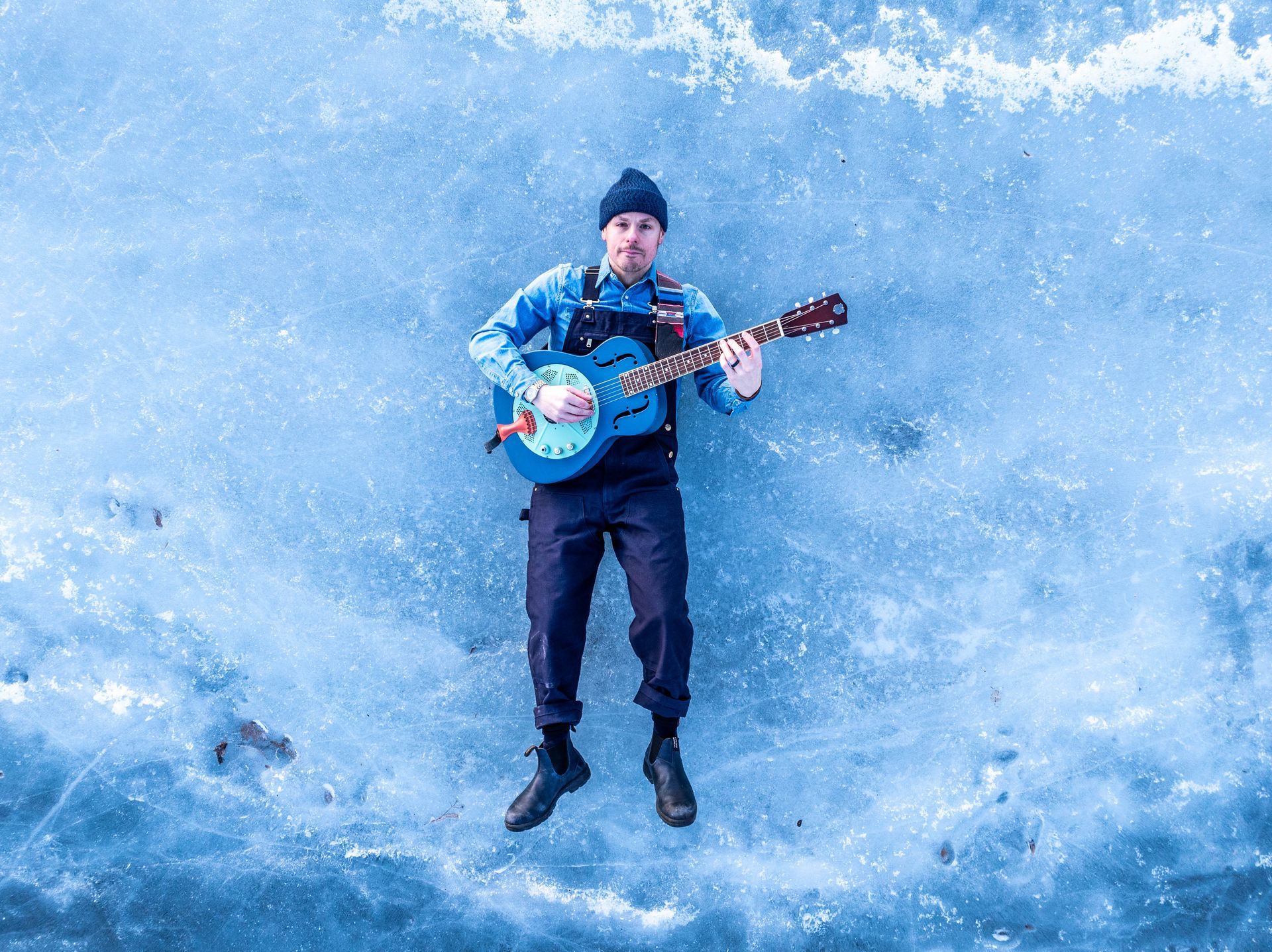 Man lying on ice, playing a blue banjo. Wearing a blue shirt, black overalls, beanie, and shoes.