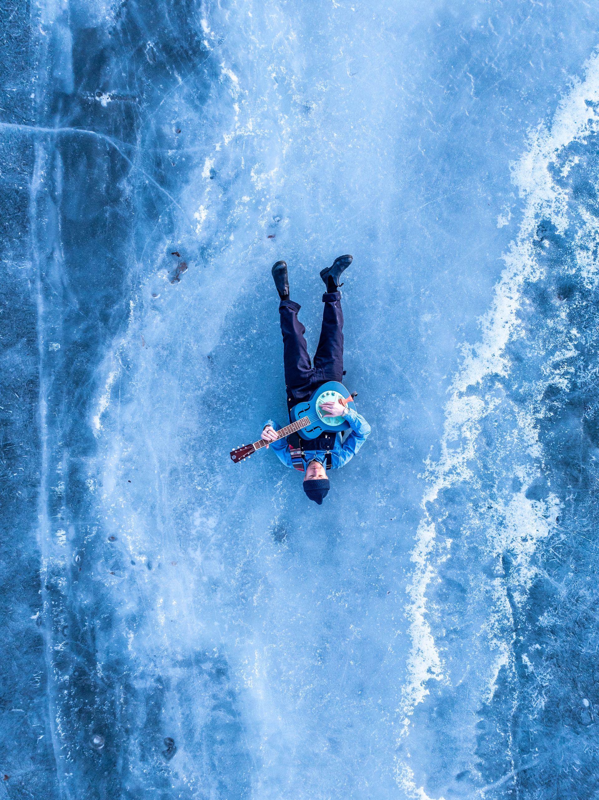 Person playing guitar on a frozen lake; overhead view. Blue tones.