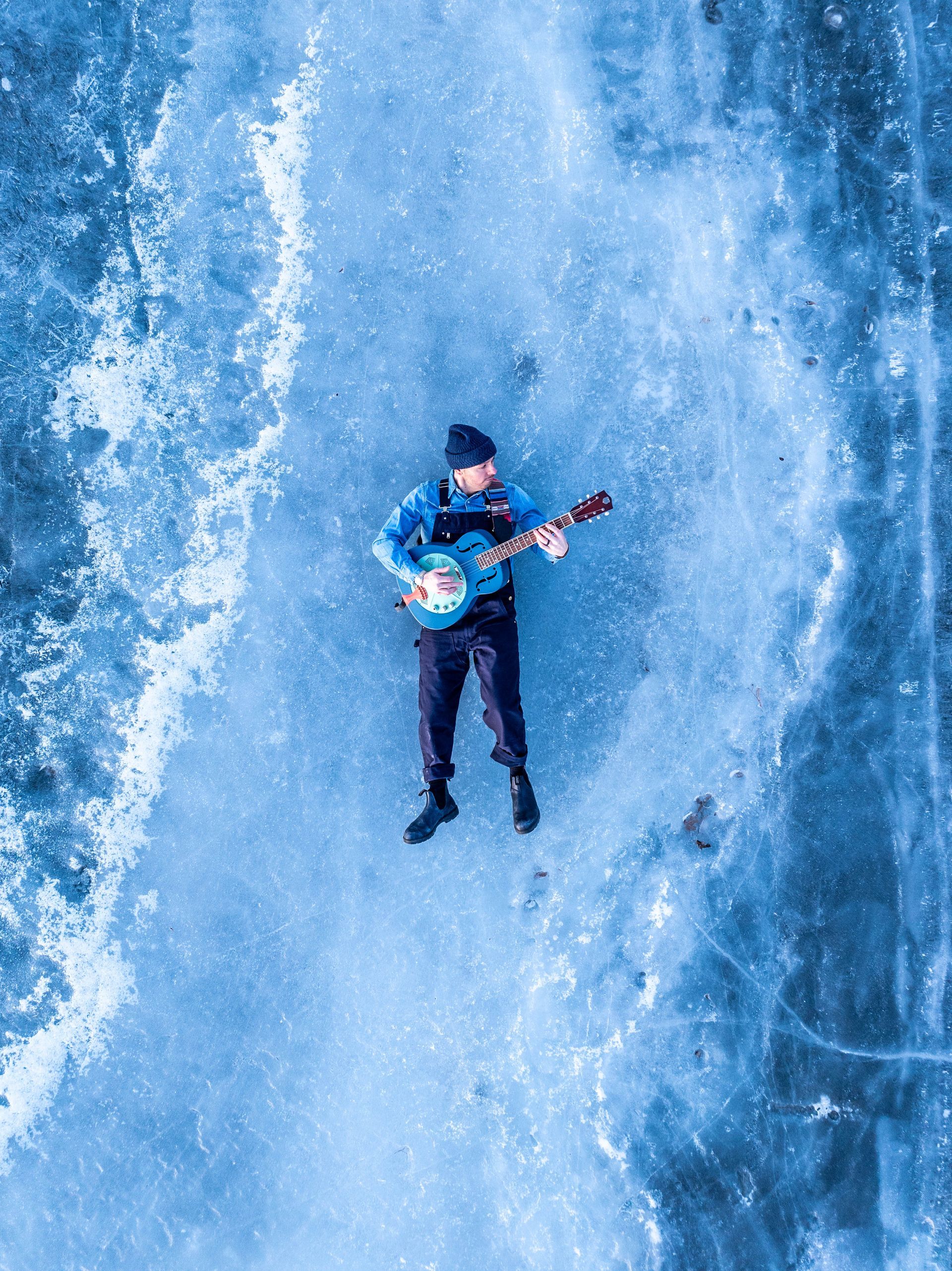 Man lying on ice, playing a guitar. Wearing blue shirt, black pants, and beanie. Overhead shot, blue tones.