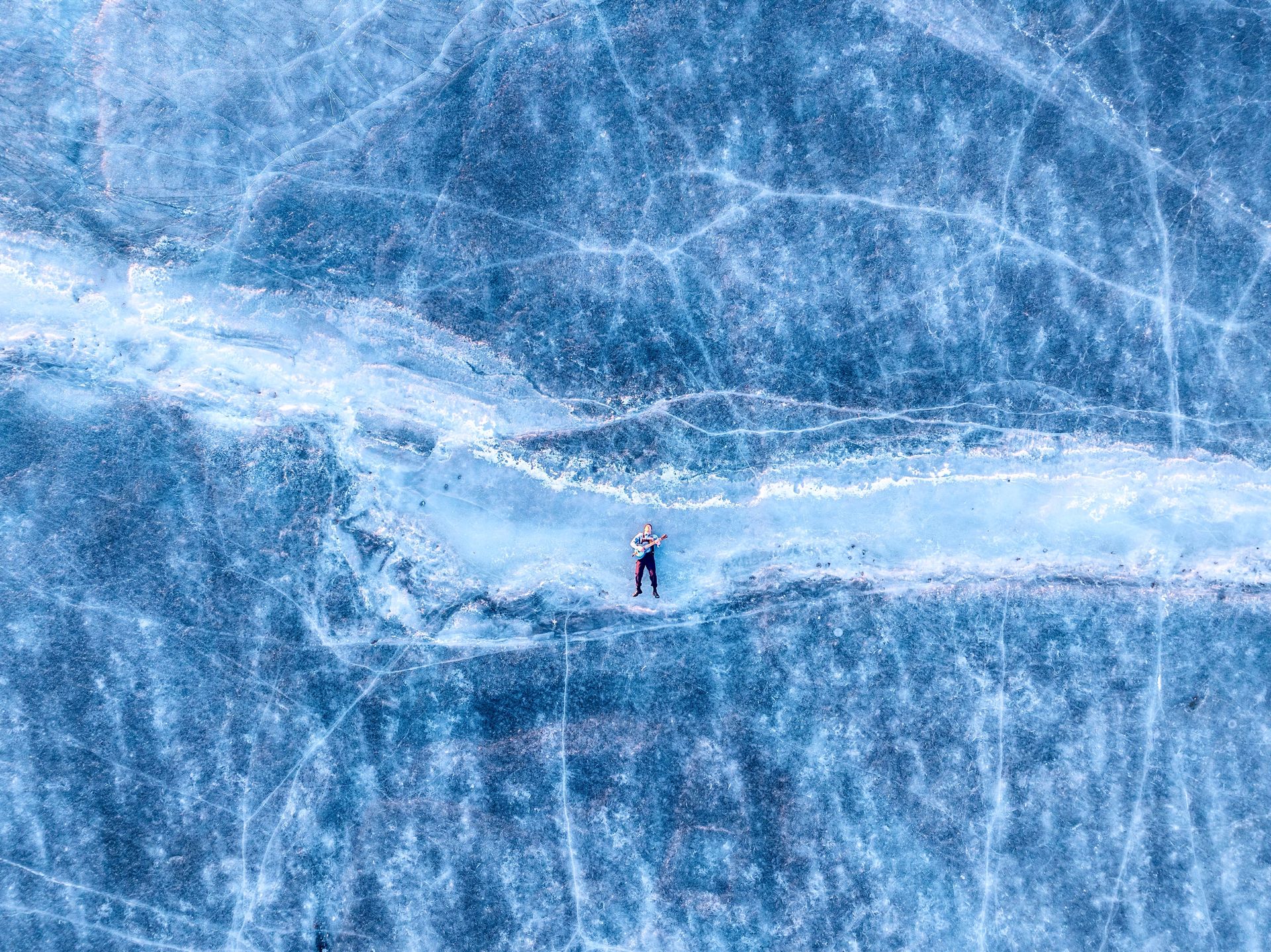 Person lying on a strip of ice, surrounded by a frozen lake with blue hues.