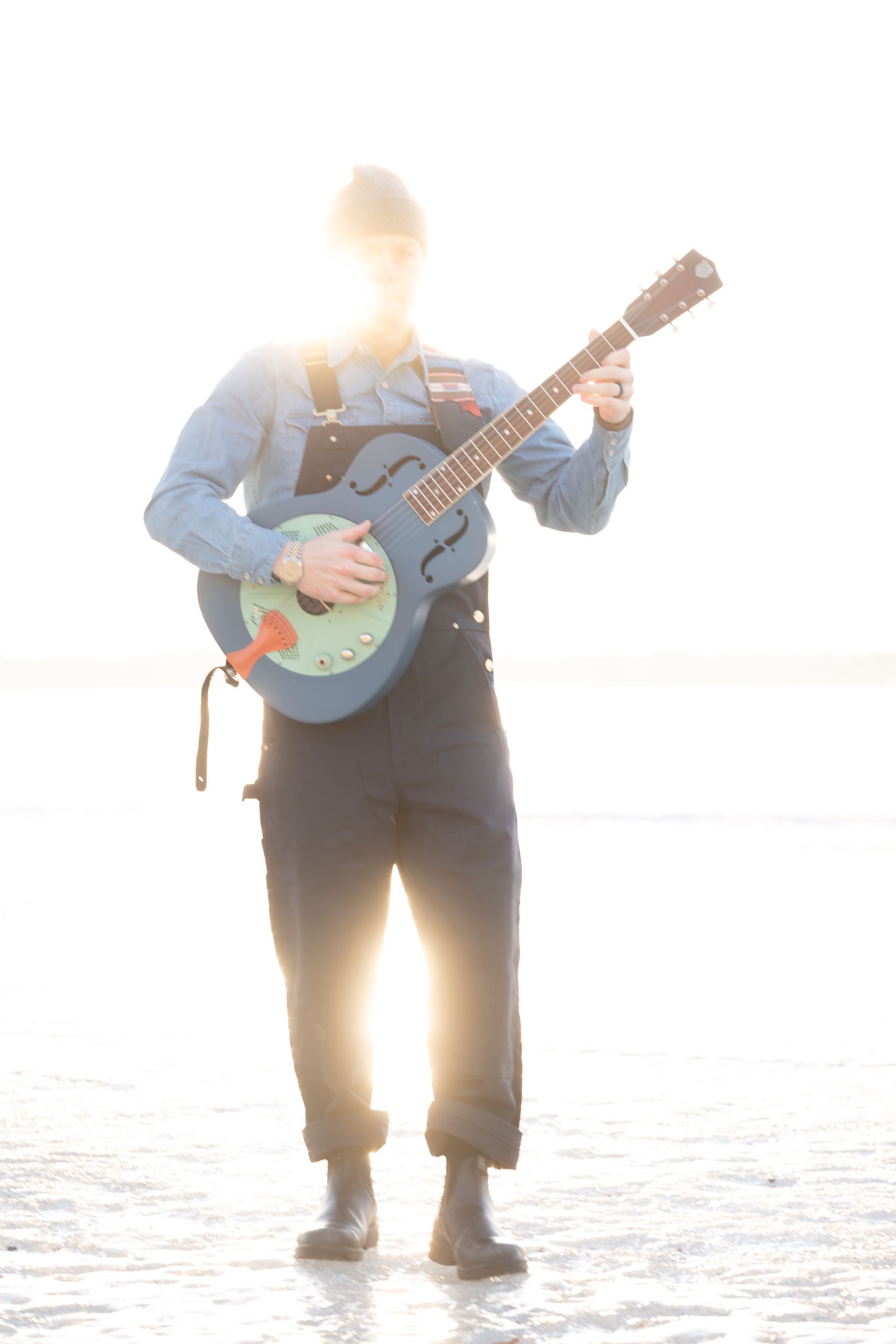 Person standing in overalls plays a blue, circular guitar with the sun behind them.