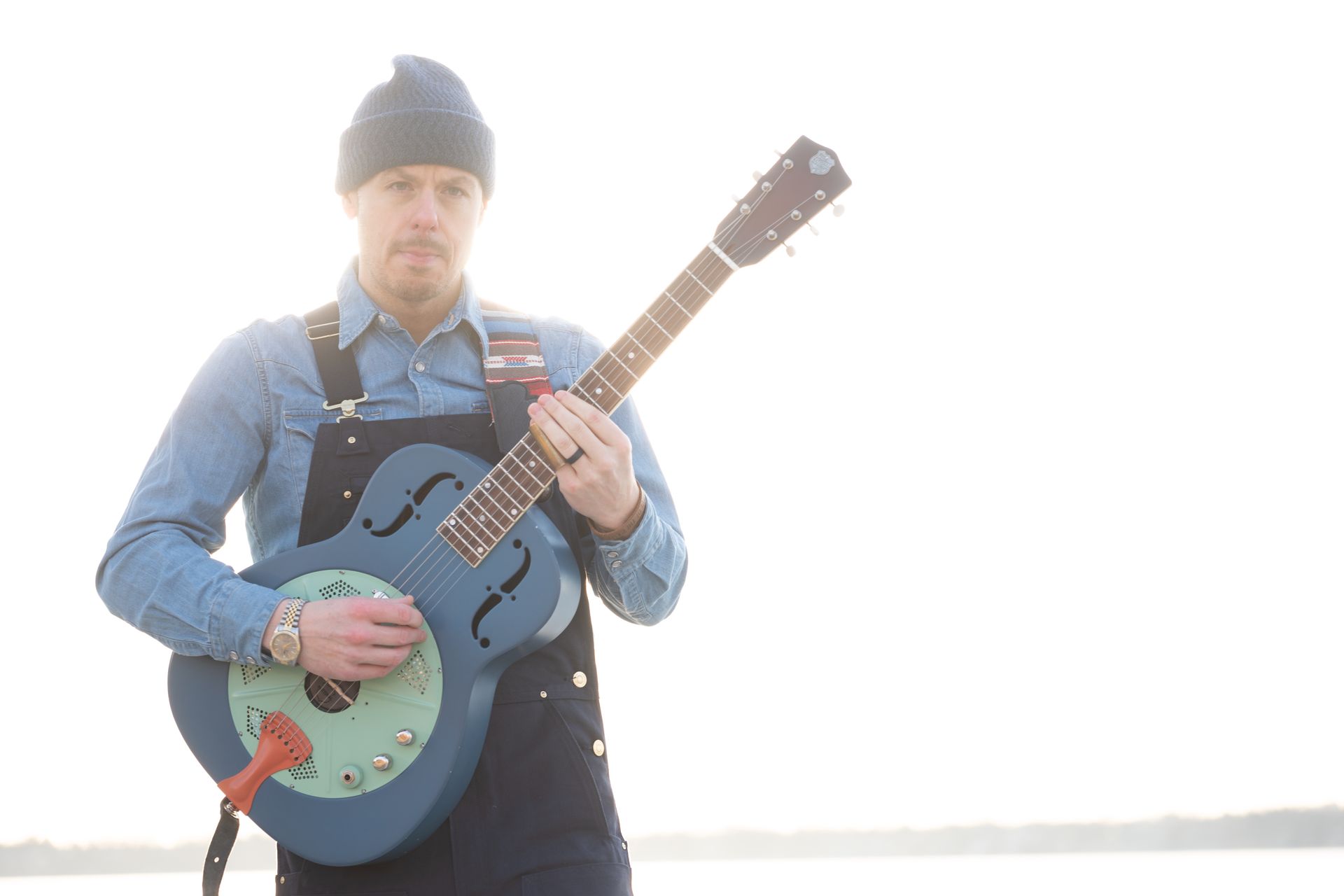 Man in overalls and beanie playing a blue guitar outdoors.