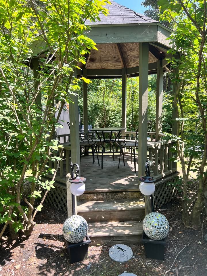 A wooden gazebo with steps, surrounded by trees. Inside are chairs and a table. Decorative globe lights and stepping stones lead to the gazebo.
