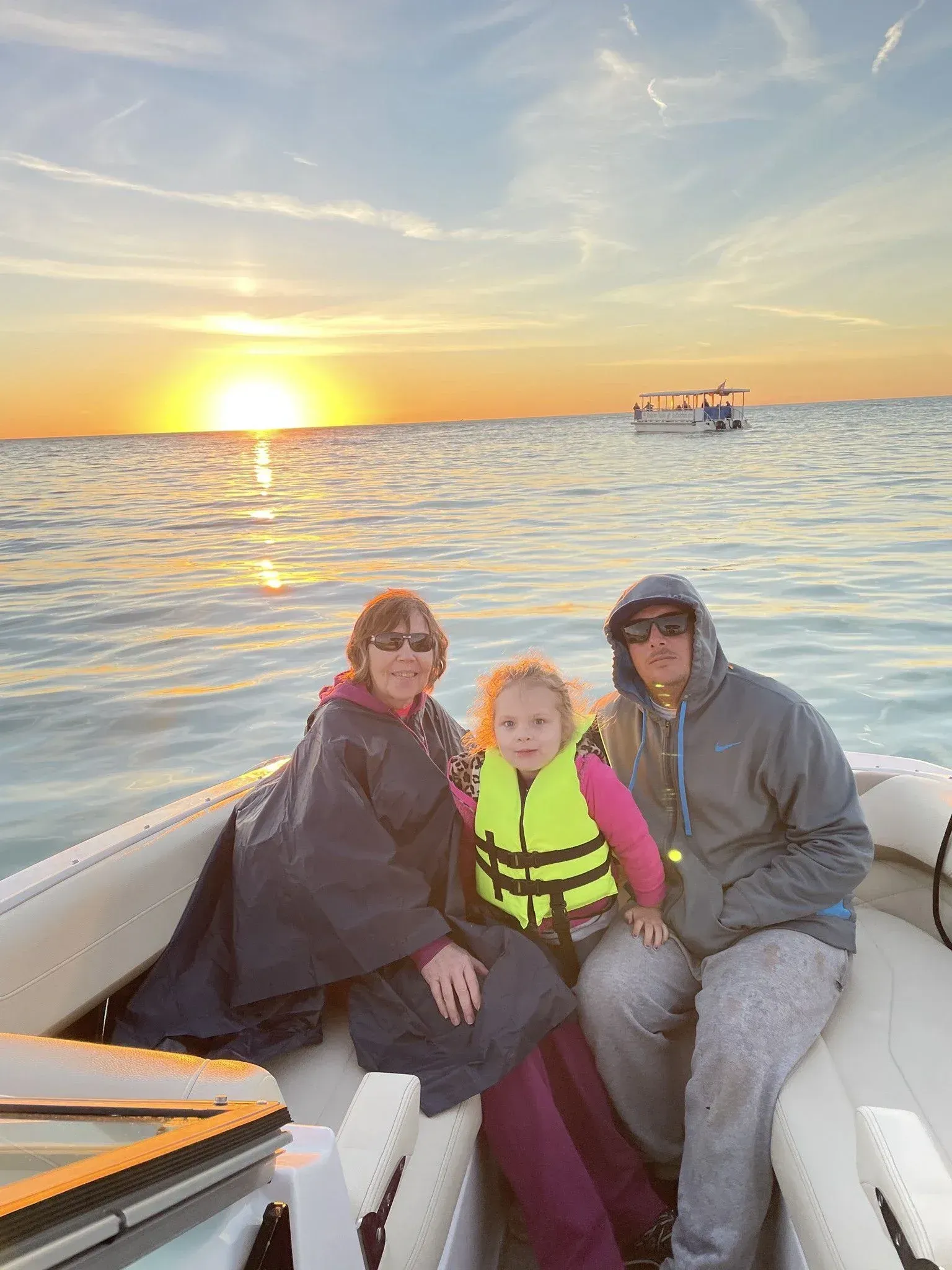 Three people sitting in a boat at sunset on calm water, with a small boat in the distance.