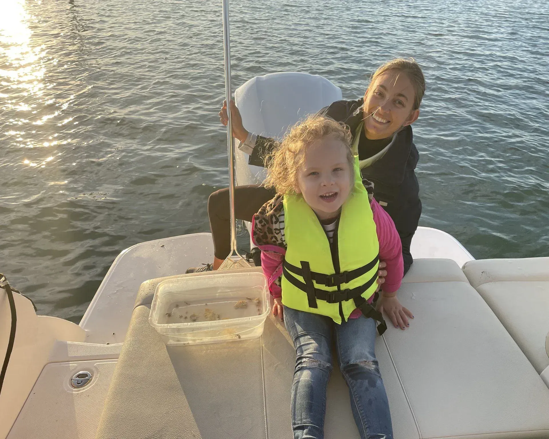 Two children smiling on a boat, wearing life jackets, with water in the background.