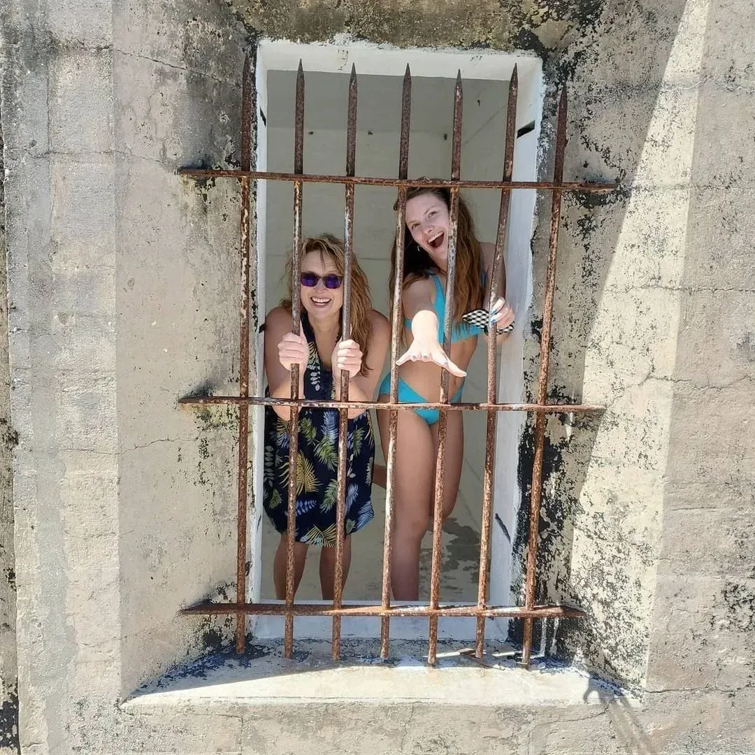Two girls smiling behind rusty bars in a window opening by a rough concrete wall.