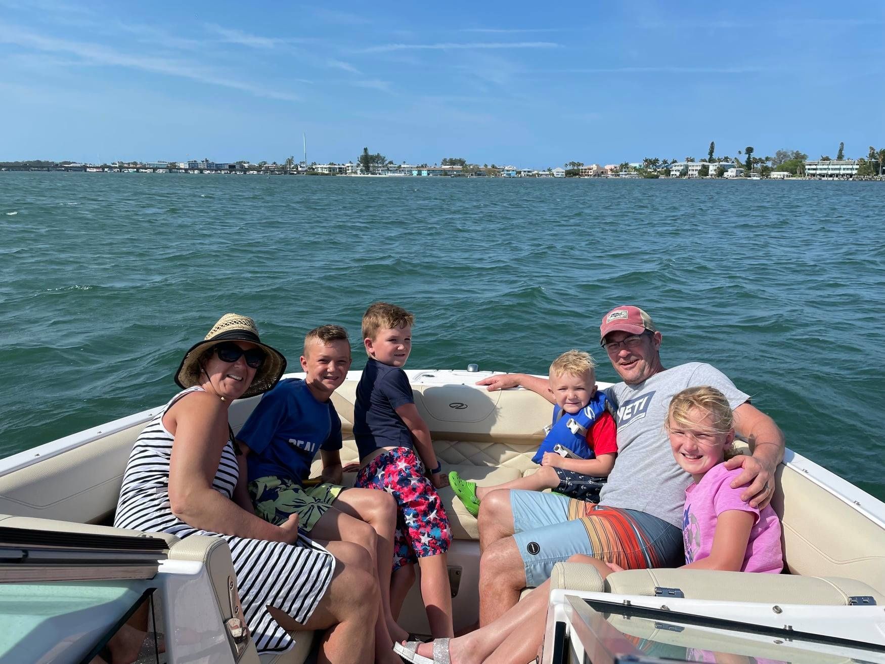 Family on a boat, smiling, enjoying sunny day on the water. Buildings visible in the background.