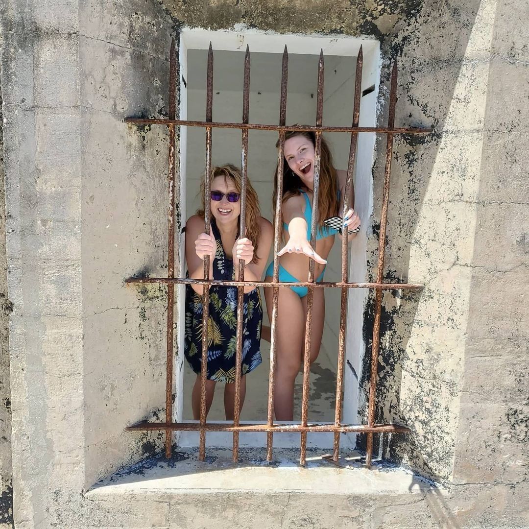 Two smiling women behind rusty bars in a weathered window. One wears a cover-up, the other a bikini, both holding the bars.