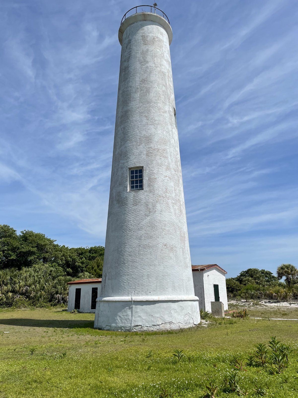 White lighthouse on a grassy field, with two small white buildings beside it. Blue sky with scattered clouds.