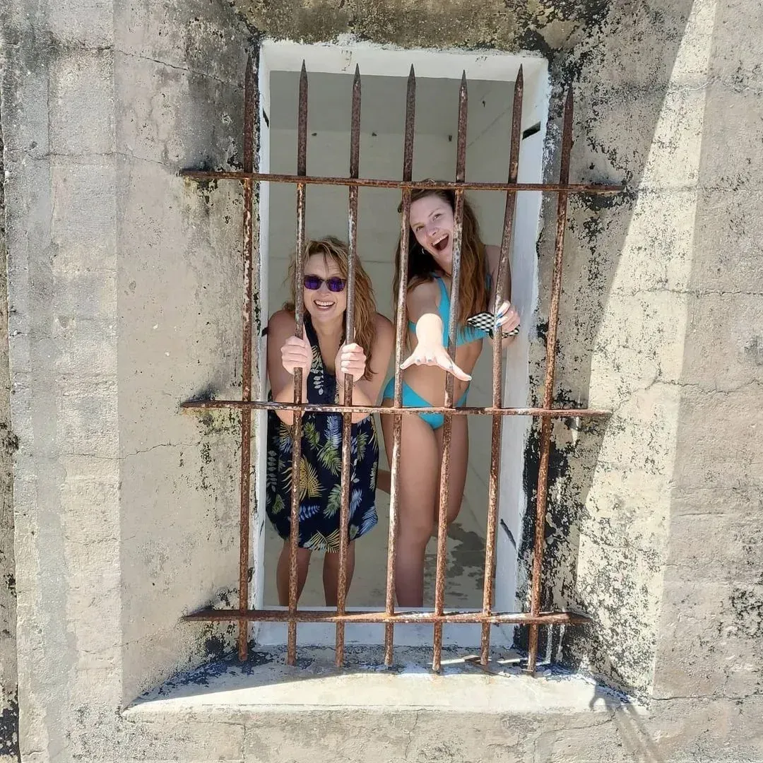 Two people standing behind rusty bars in a concrete window opening, smiling at the camera.