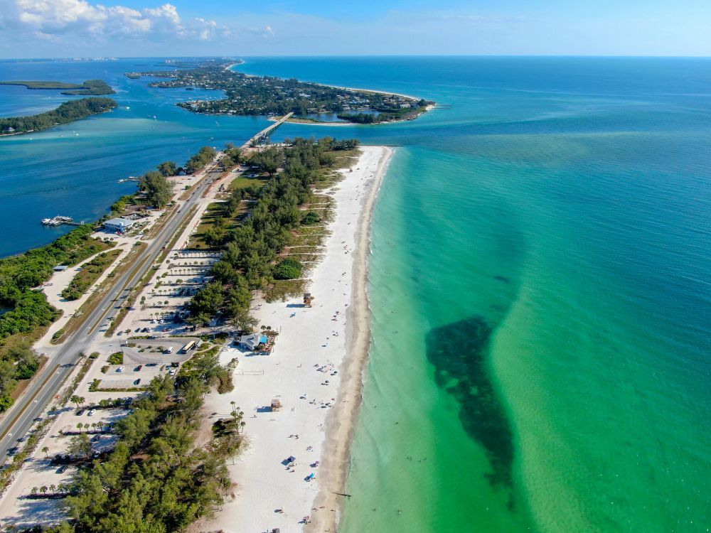 Aerial view of a white sandy beach with turquoise water. A road and bridge connect it to a nearby island under a blue sky.