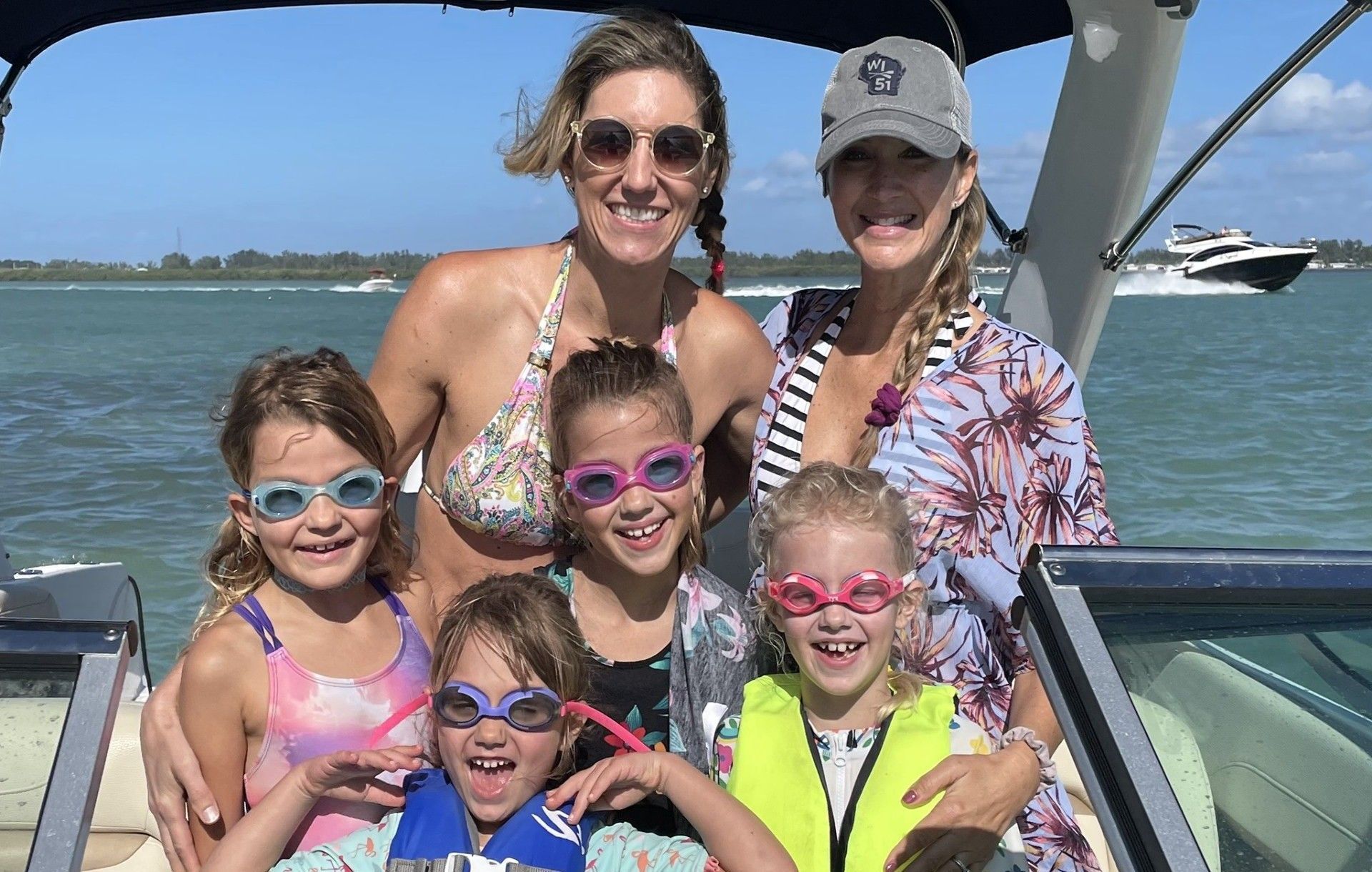 Two women and four young girls on a boat, smiling, wearing swimwear and goggles on a sunny day.