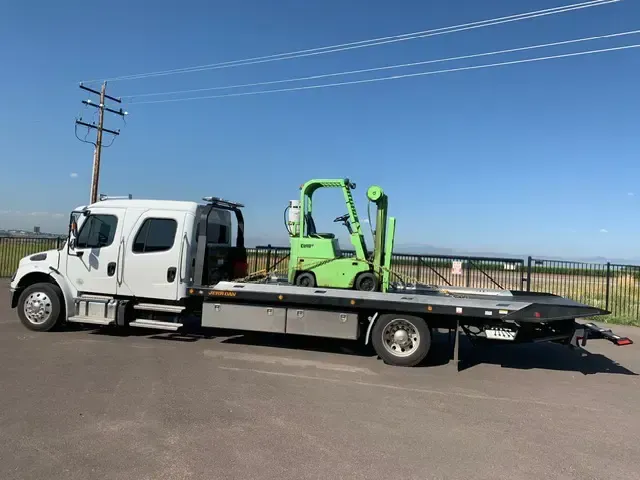 White flatbed tow truck with a green forklift on the bed, parked on a road.
