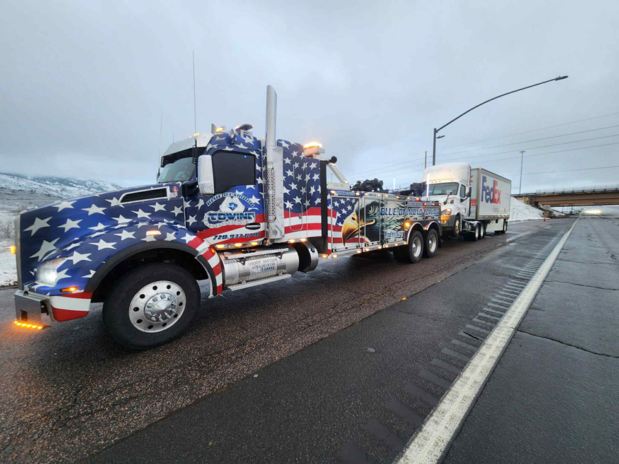 Tow truck with American flag design towing a FedEx truck on a road.
