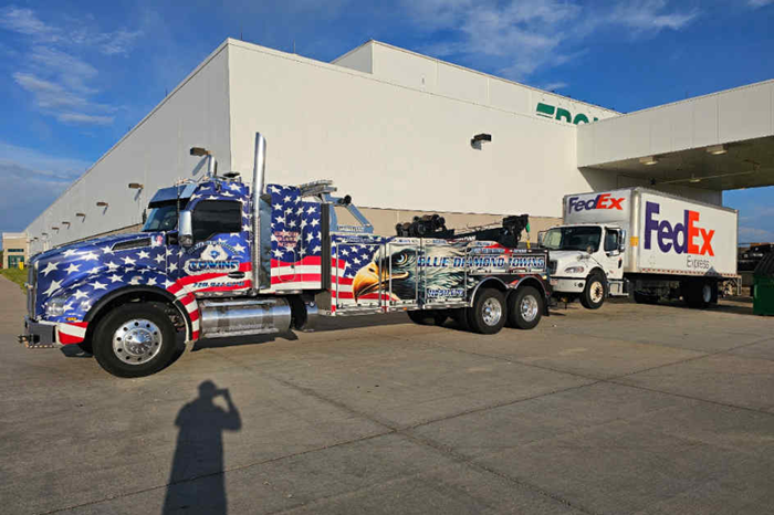 Tow truck with American flag design towing a FedEx truck in front of a FedEx building.