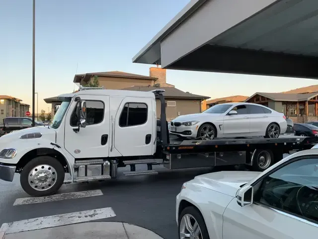 White tow truck carrying a white BMW car; parked near a building in daytime.
