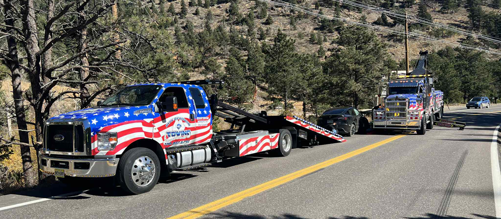 Two tow trucks with American flag paint jobs on a roadside, towing a car.