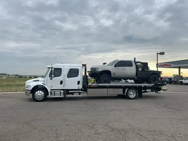 White tow truck carrying a light-colored pickup truck on a flatbed. Outdoors, cloudy sky, gas station in the distance.