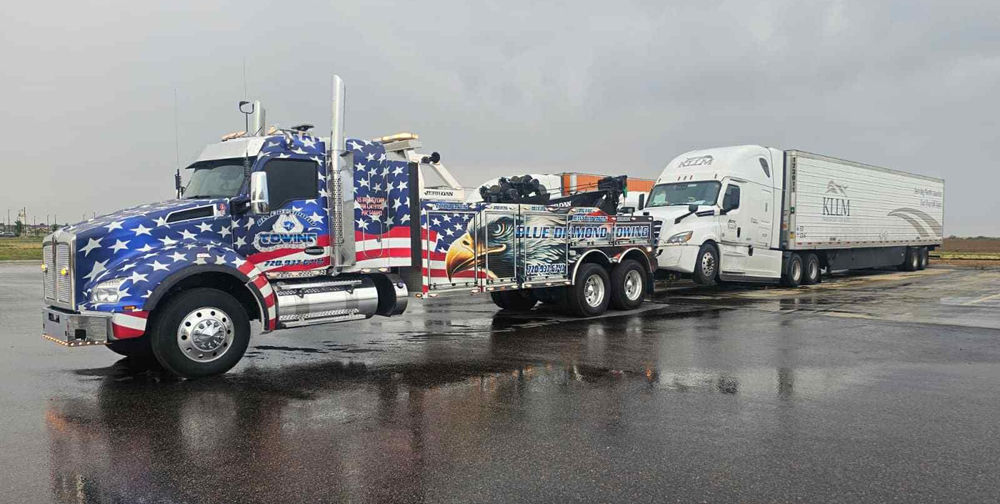 Tow truck with American flag design towing a semi-truck and trailer on a wet, paved surface.