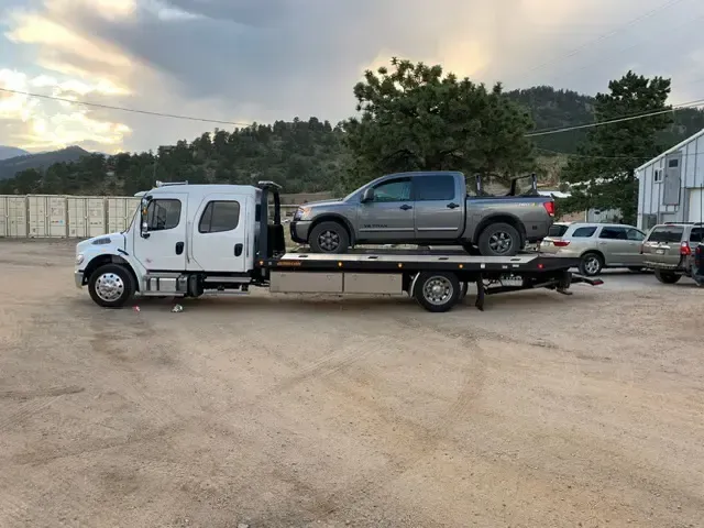 Tow truck with gray pickup truck loaded, in front of a building and trees.