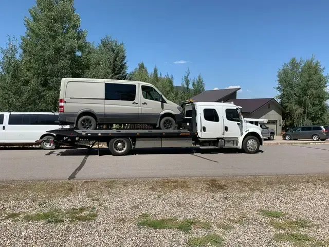 A gray van being towed on a flatbed truck on a sunny road, next to a white limousine.