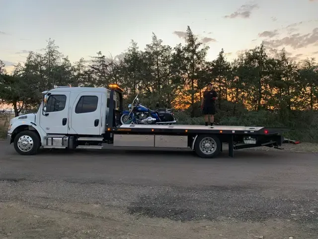 White tow truck with motorcycle on flatbed, parked on road at sunset.