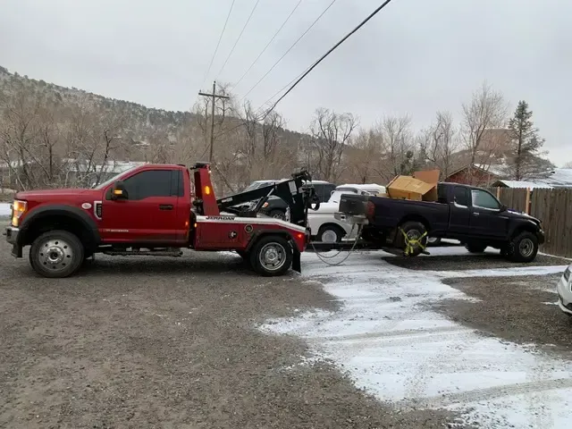 Red tow truck towing a black pickup truck on a snowy, gravel lot.