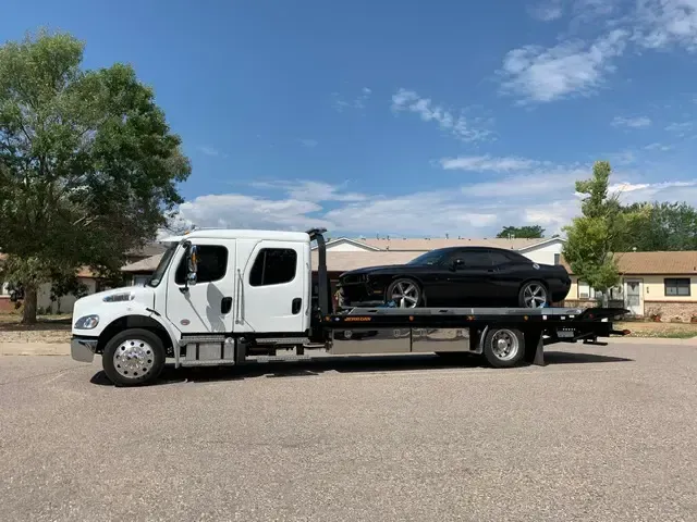 A black car on a white tow truck on a residential street under a blue sky.