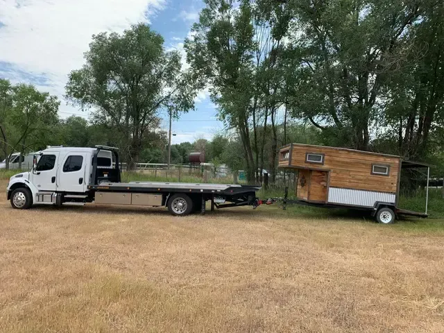 A white flatbed tow truck pulling a tiny wooden house trailer on a grassy field.
