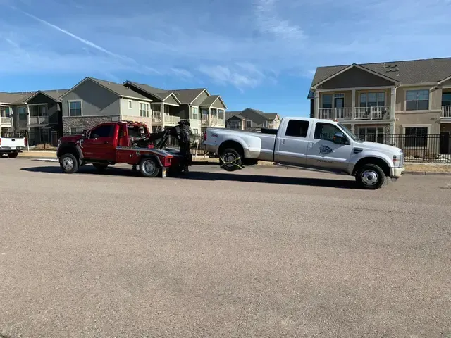A red tow truck towing a white pickup truck on a paved road in front of apartment buildings.