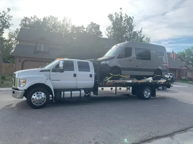White tow truck hauling a gray camper van on a residential street.