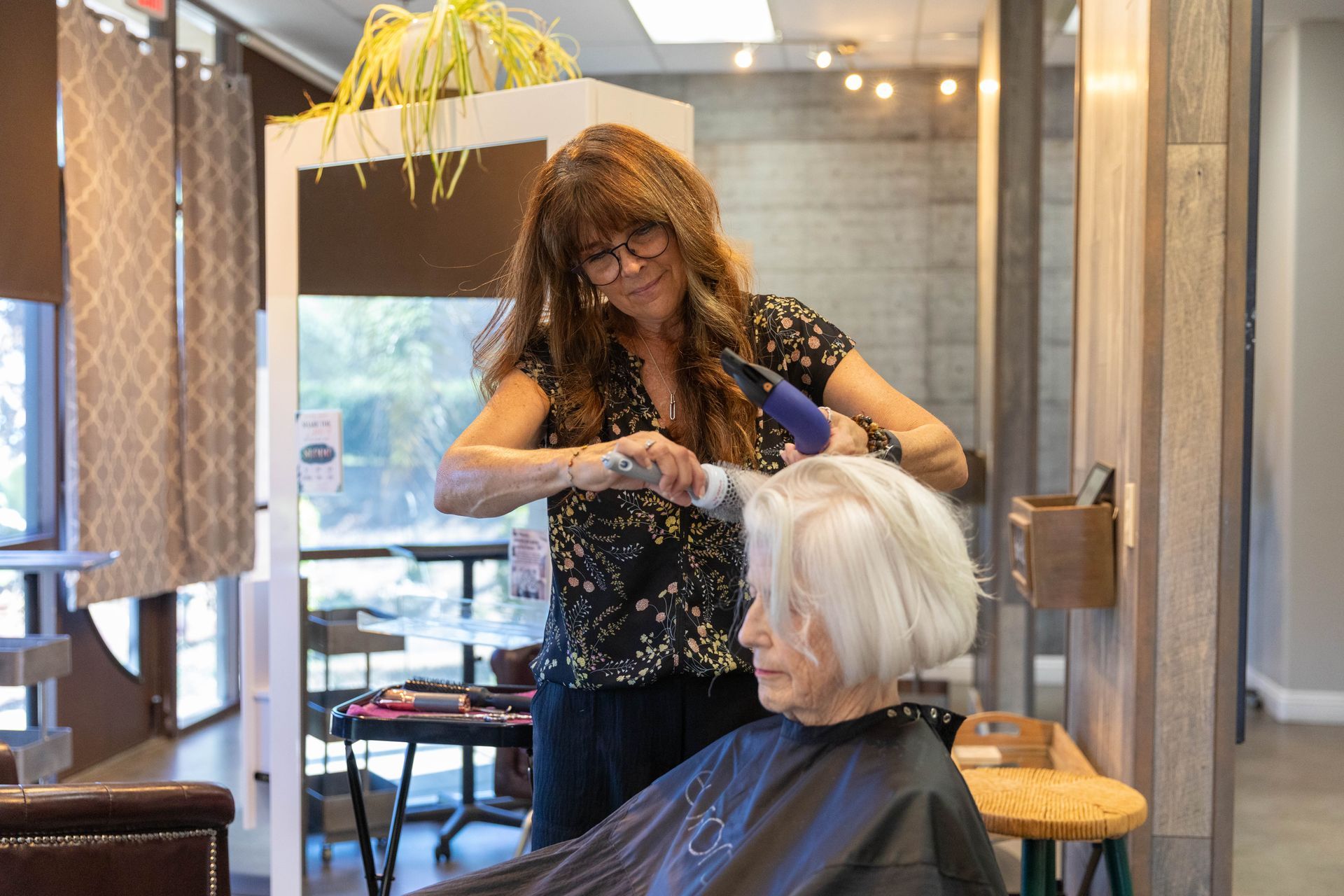 A stylist using a hairdryer on a client's red hair in a salon. The client smiles.