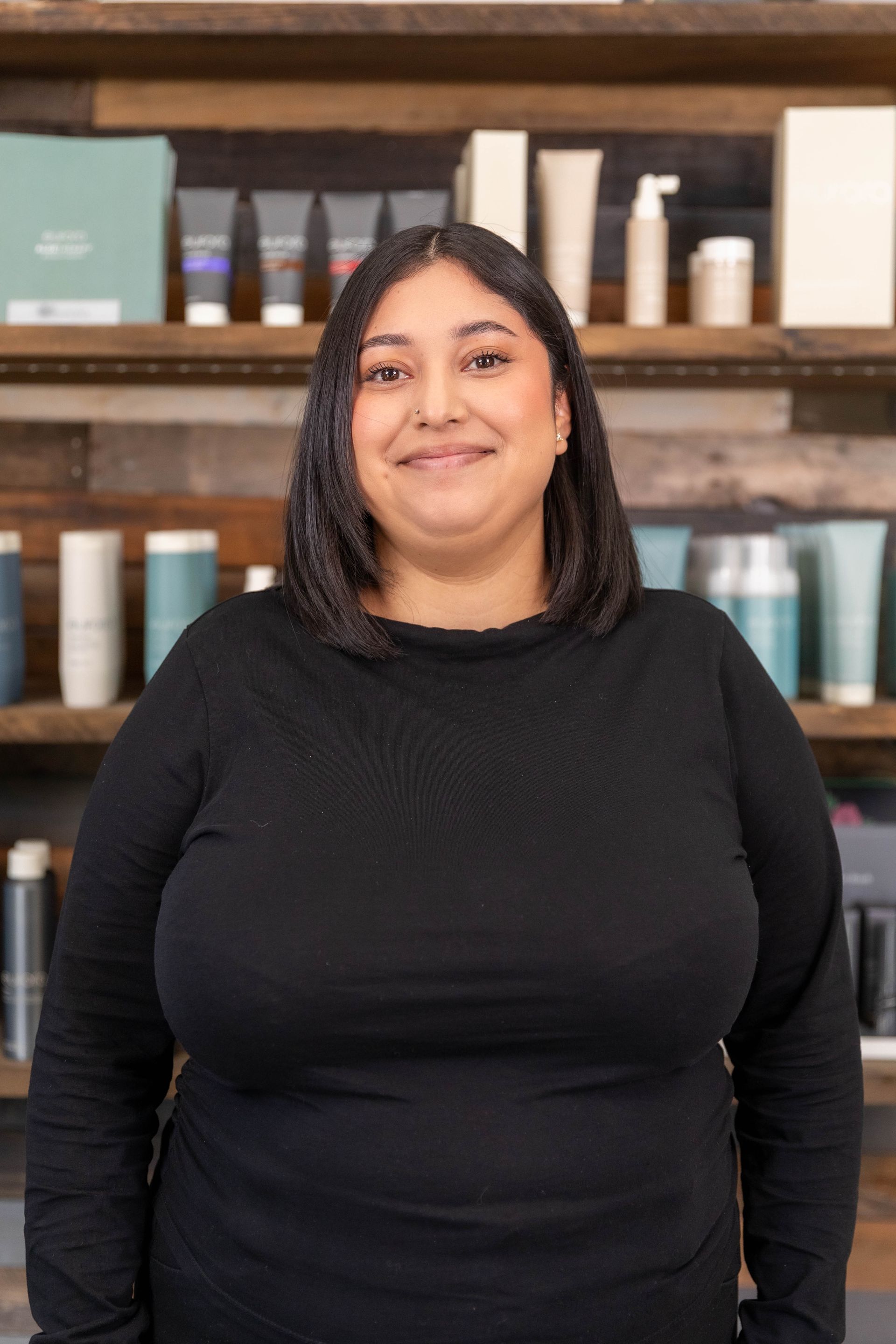 Smiling person in black top standing indoors before shelves of hair care products