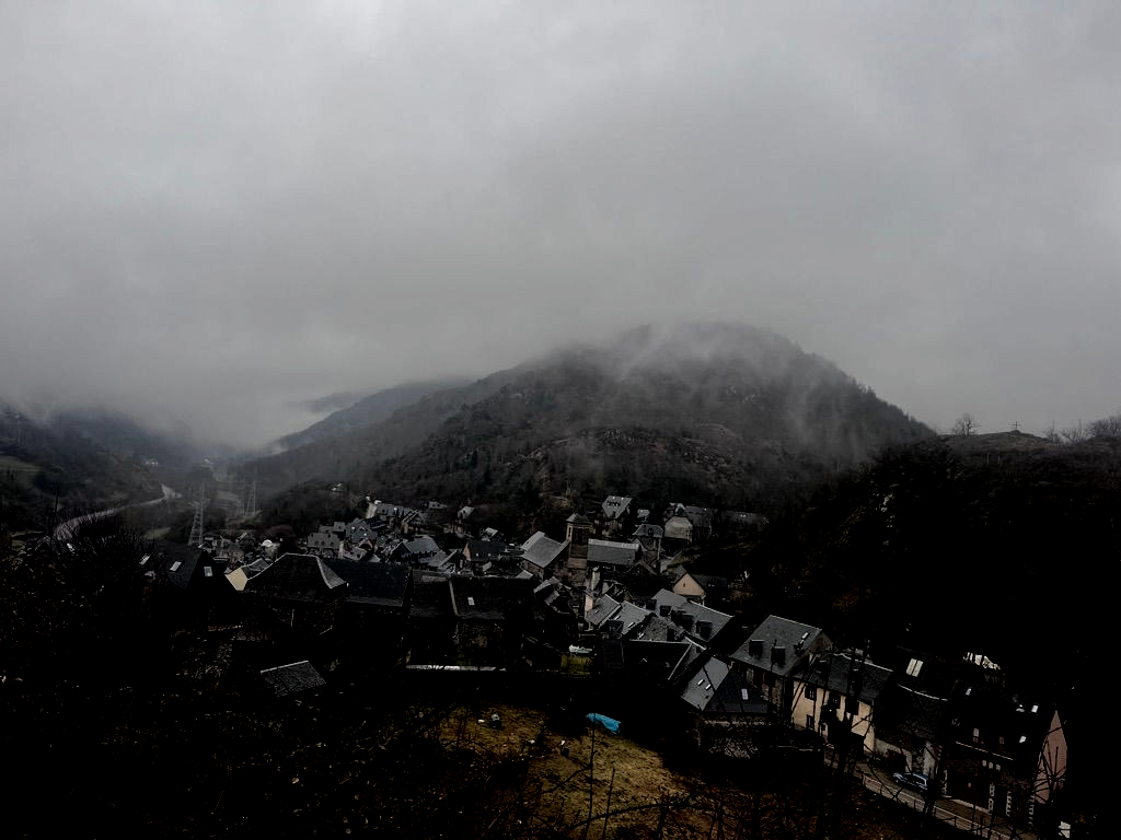 Pueblo brumoso en la ladera de una colina, con tejados grises, bajo una montaña cubierta de niebla.