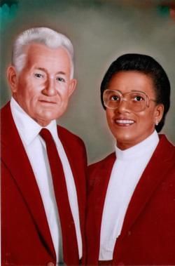 Couple in matching red suits, smiling. Man has gray hair, white tie. Woman wears glasses.