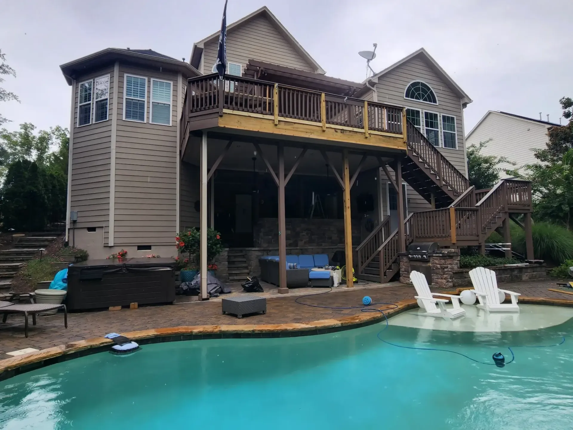 A multi-level deck and house overlook a swimming pool. Wooden deck with steps, columns, and a sitting area. White Adirondack chairs near the pool.