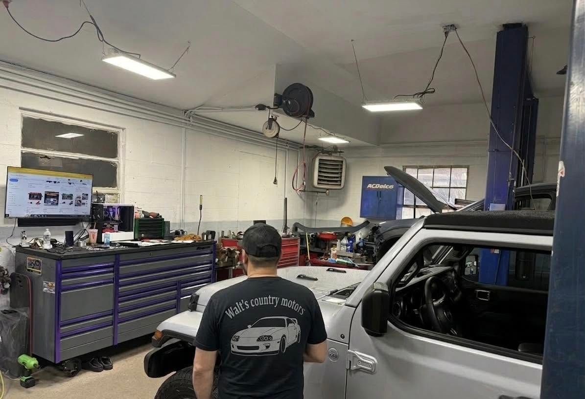 Auto repair shop with a mechanic working on a white SUV beside tool cabinets and overhead lights