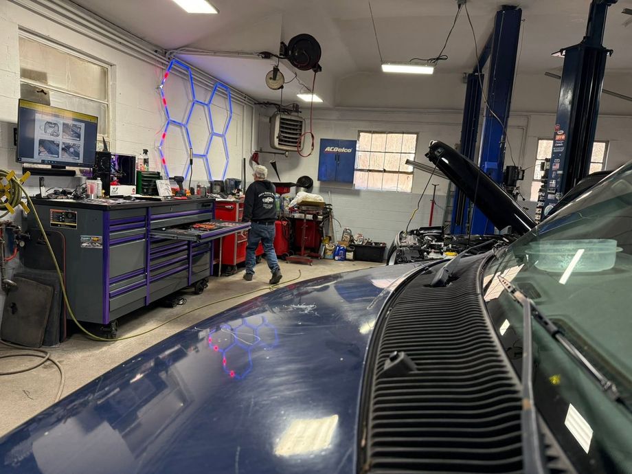 Auto repair garage with a blue car, raised hood, and mechanic working near tools and equipment.