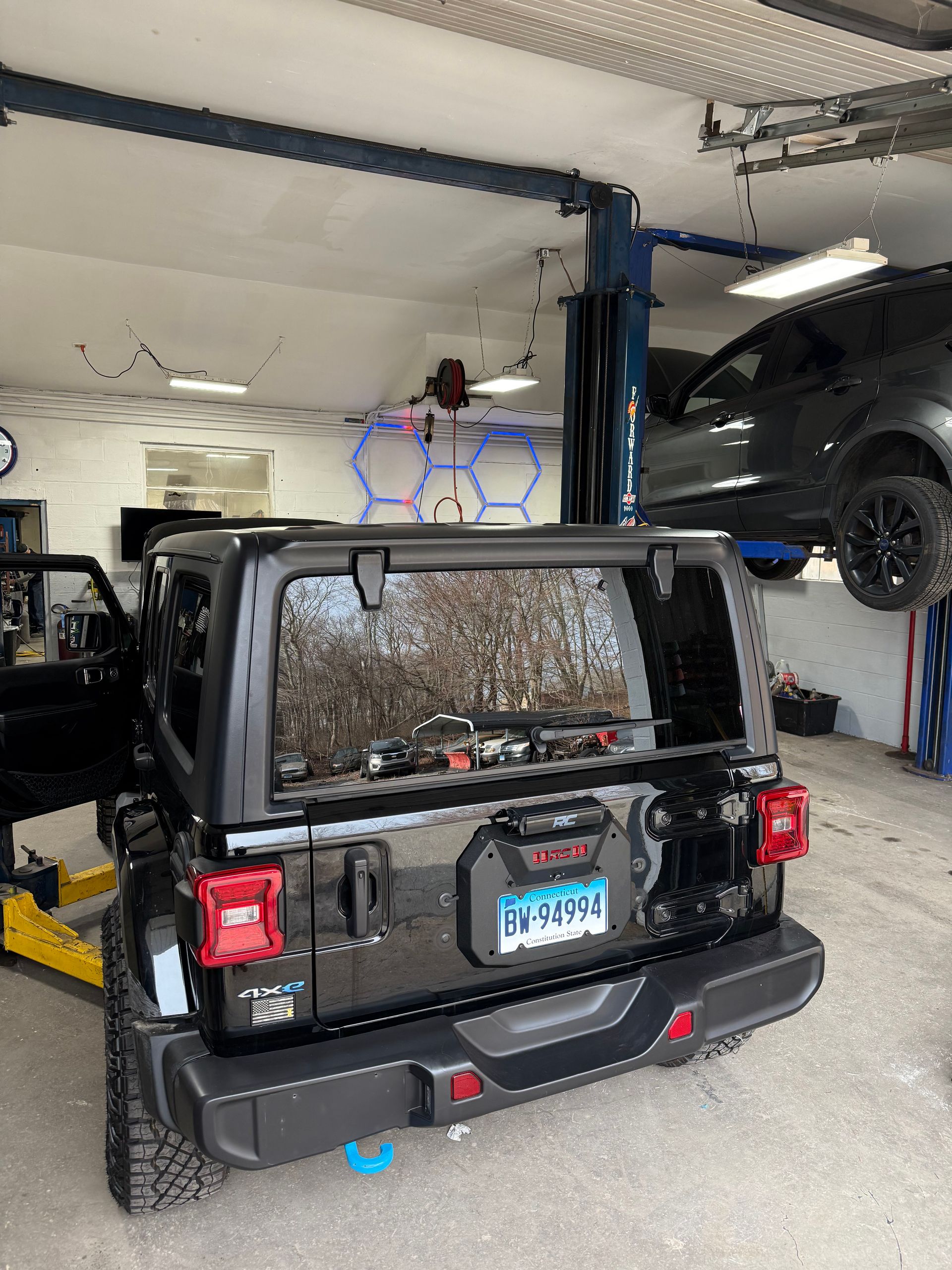 Black Jeep in a garage on a lift, with another vehicle elevated beside it.