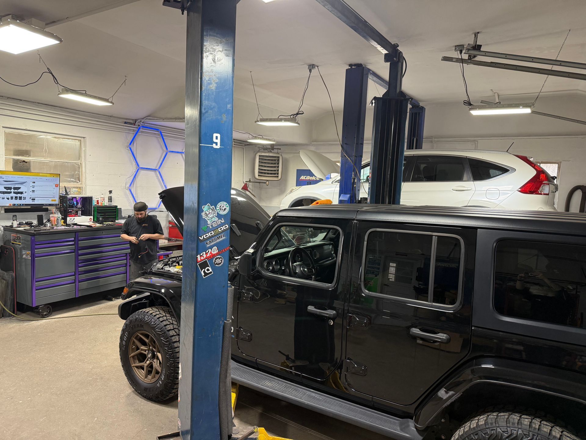 Black Jeep in auto repair shop with mechanic working nearby and a white car on a lift