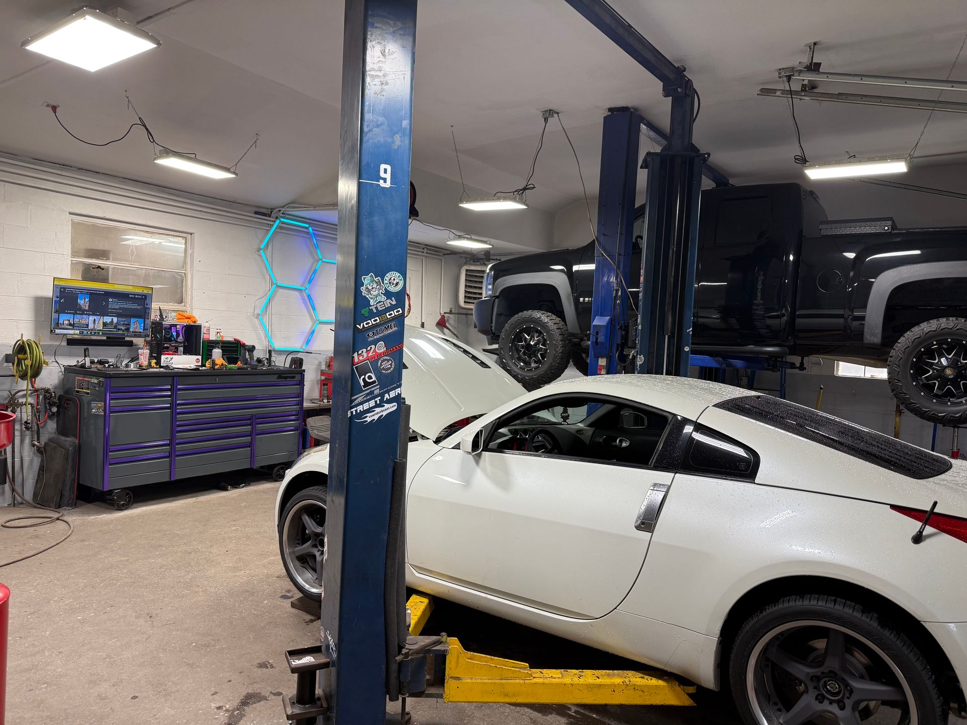 White sports car in an auto repair shop on a lift, with a black SUV and tool carts in the background
