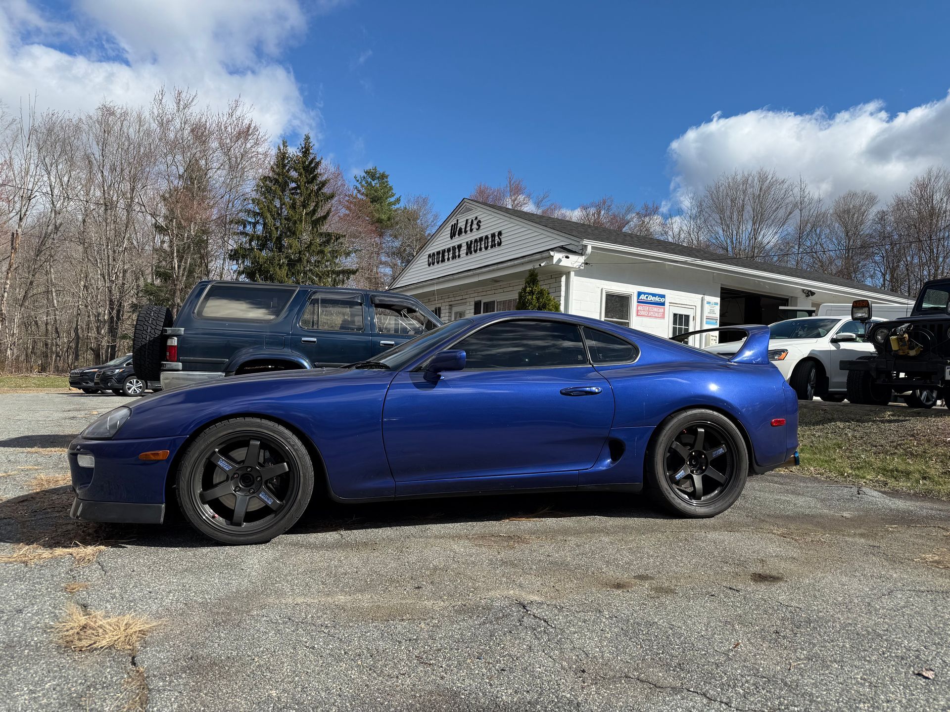 Blue sports car parked outside a building on a gravel lot with other vehicles nearby