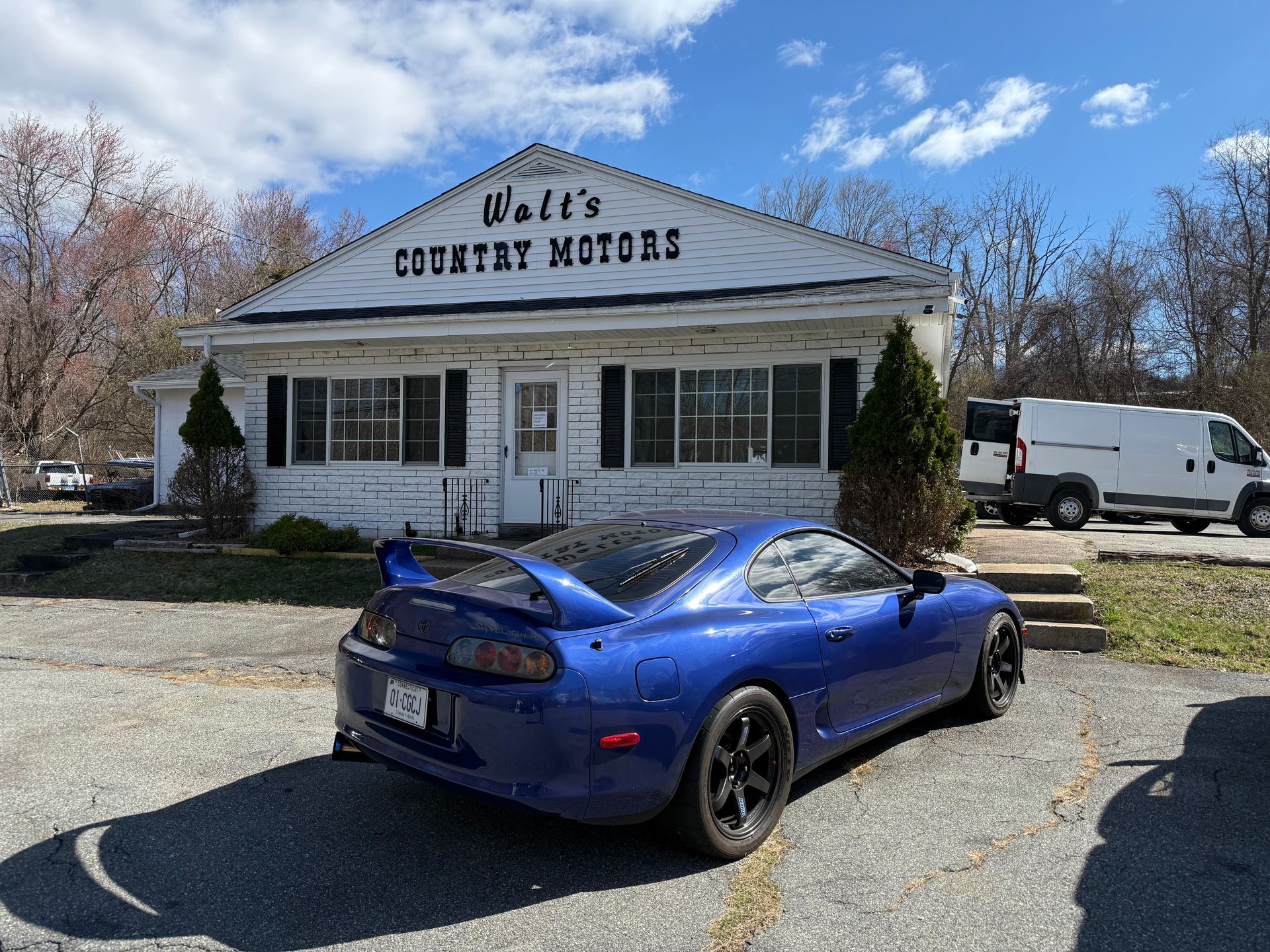 Blue sports car parked outside a white country store with a “Country Hoods” sign.
