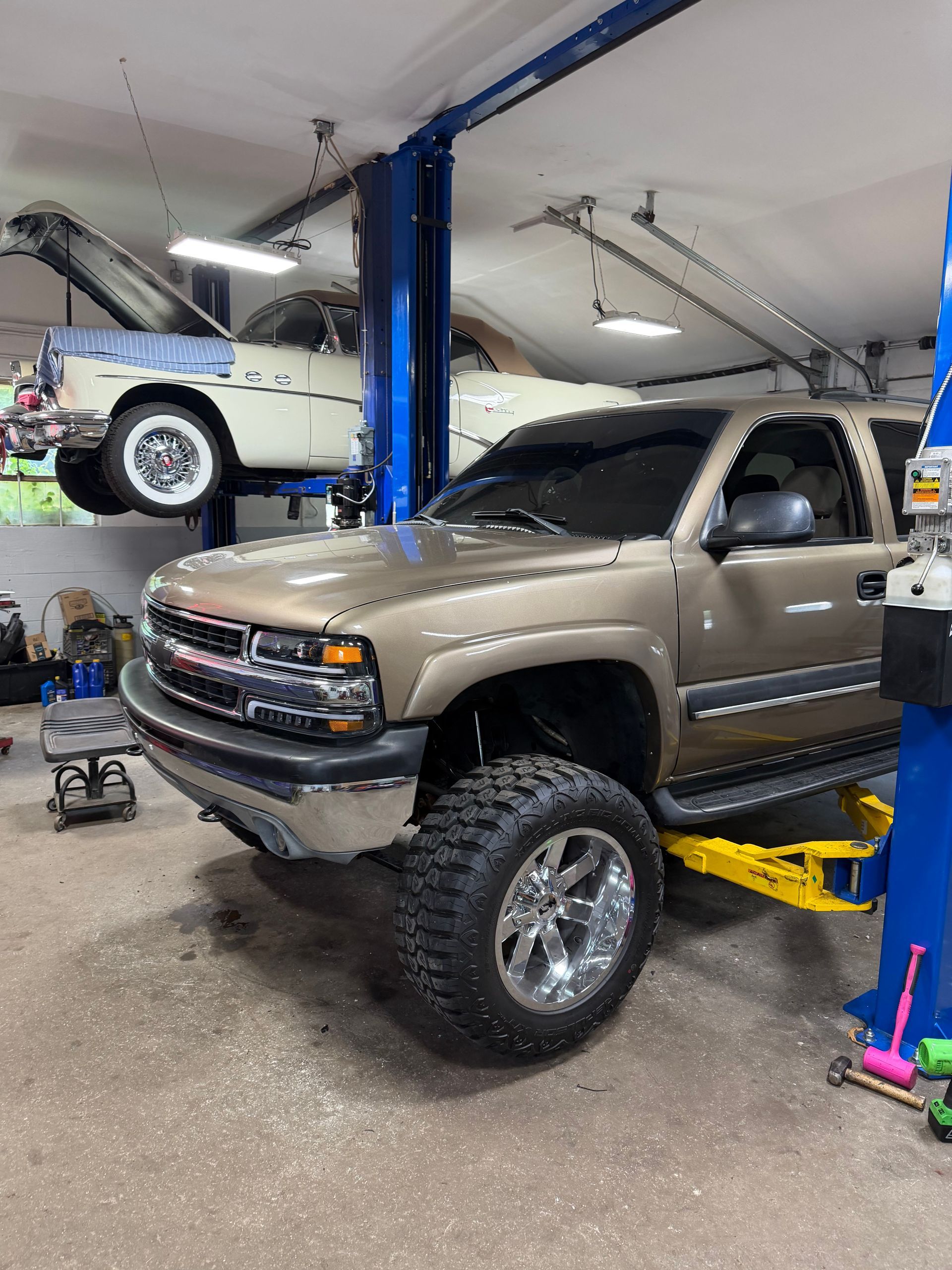 Two lifted trucks in a garage, one beige truck on a lift and one white truck suspended above.