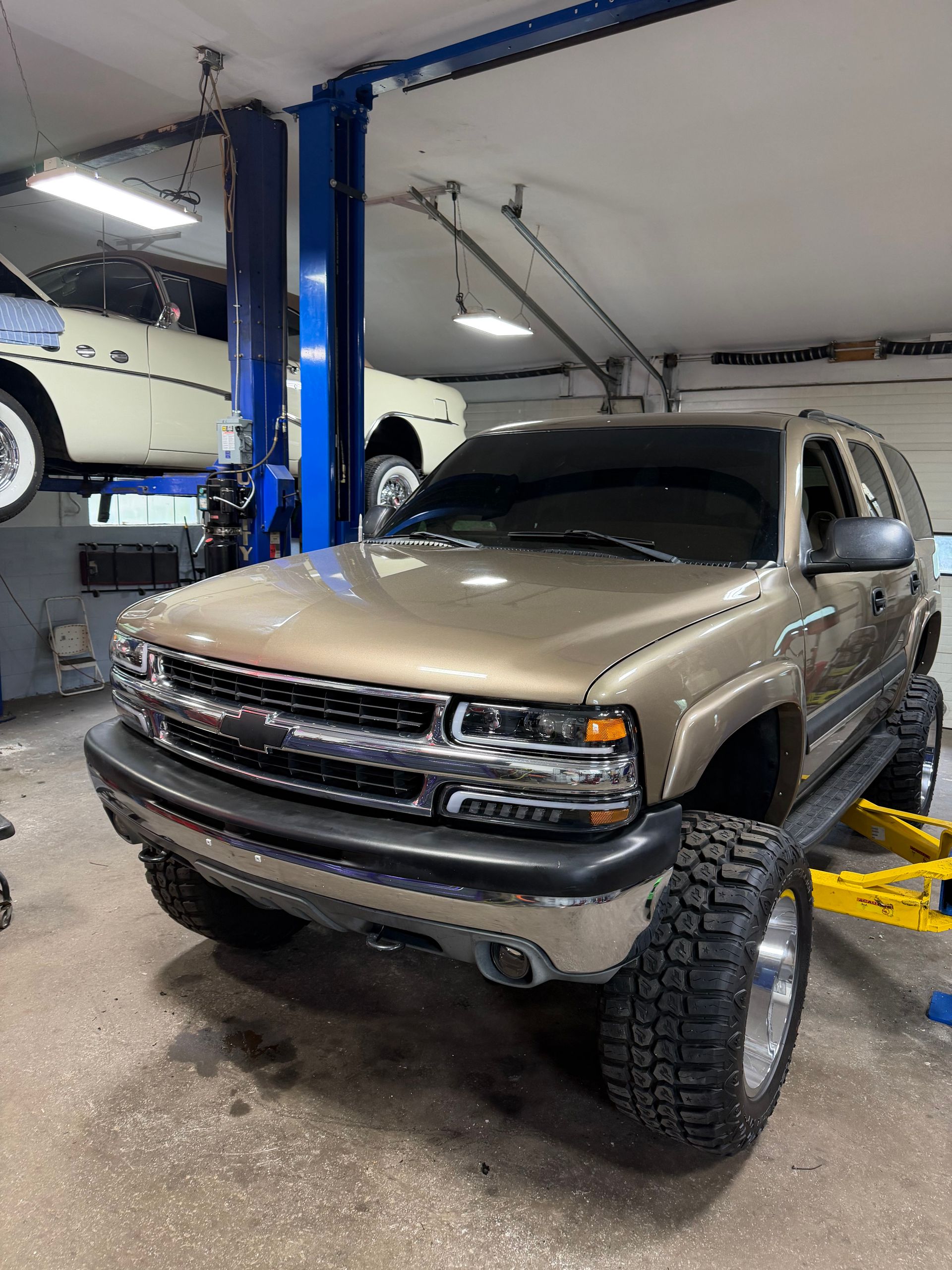 Lifted tan SUV in an auto shop garage on a blue lift, with a white car parked behind it.