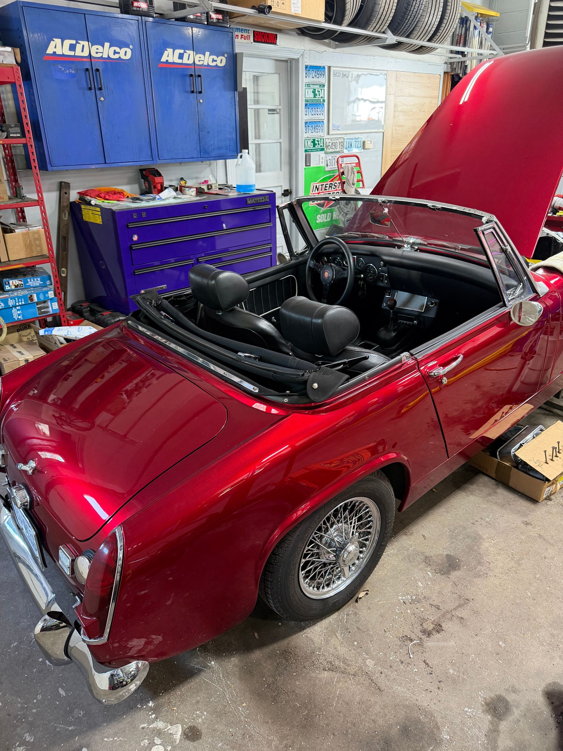 Red classic convertible in a garage, hood open, with blue tool cabinets and shop equipment nearby