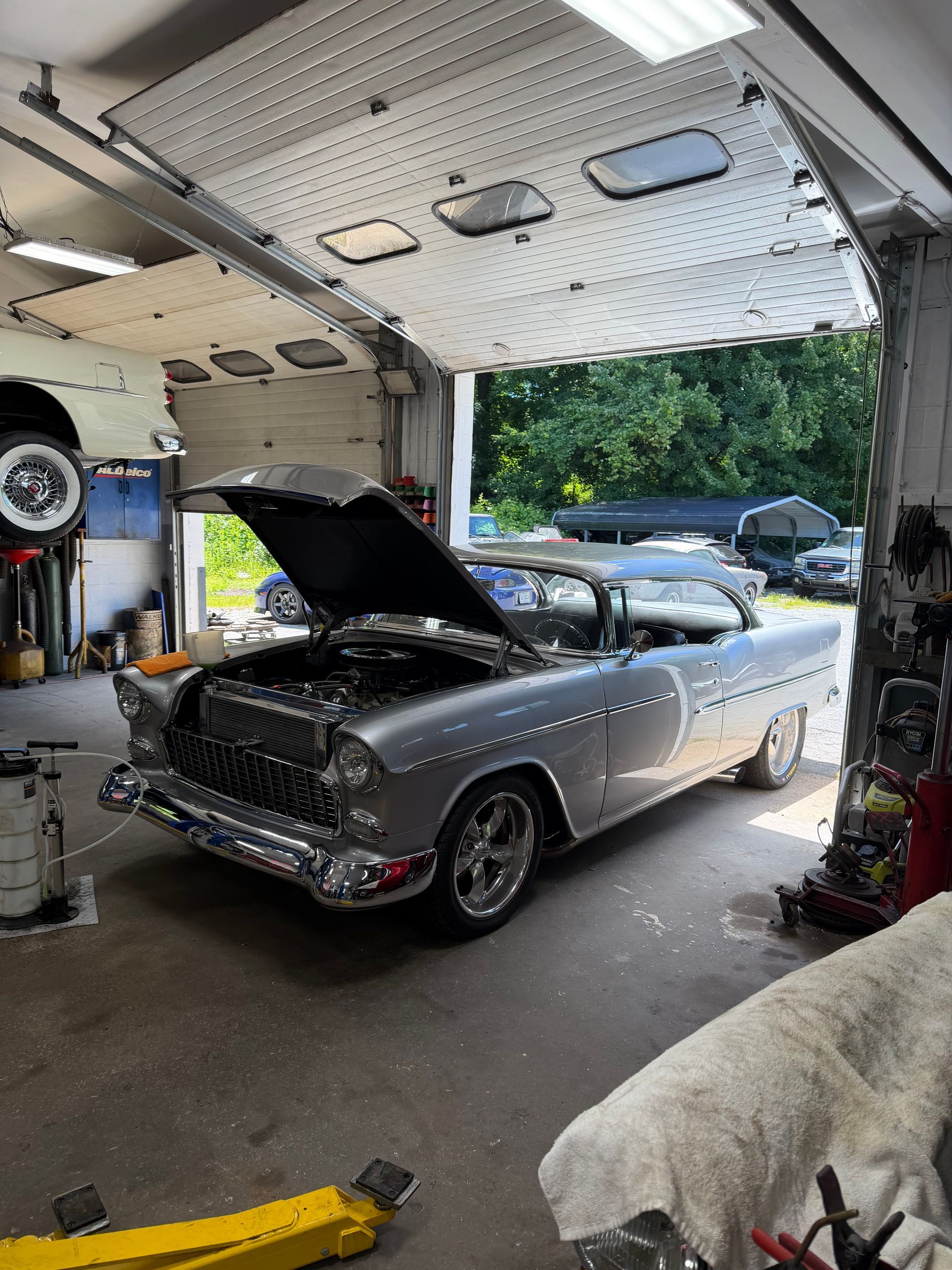 Classic silver car with hood open in a garage, showing its rear and side under bright indoor light.